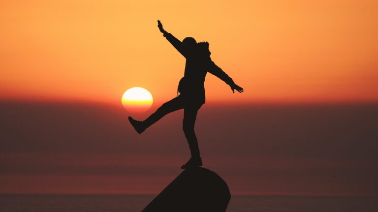 Man balancing on a rock in front of sunset with hands up